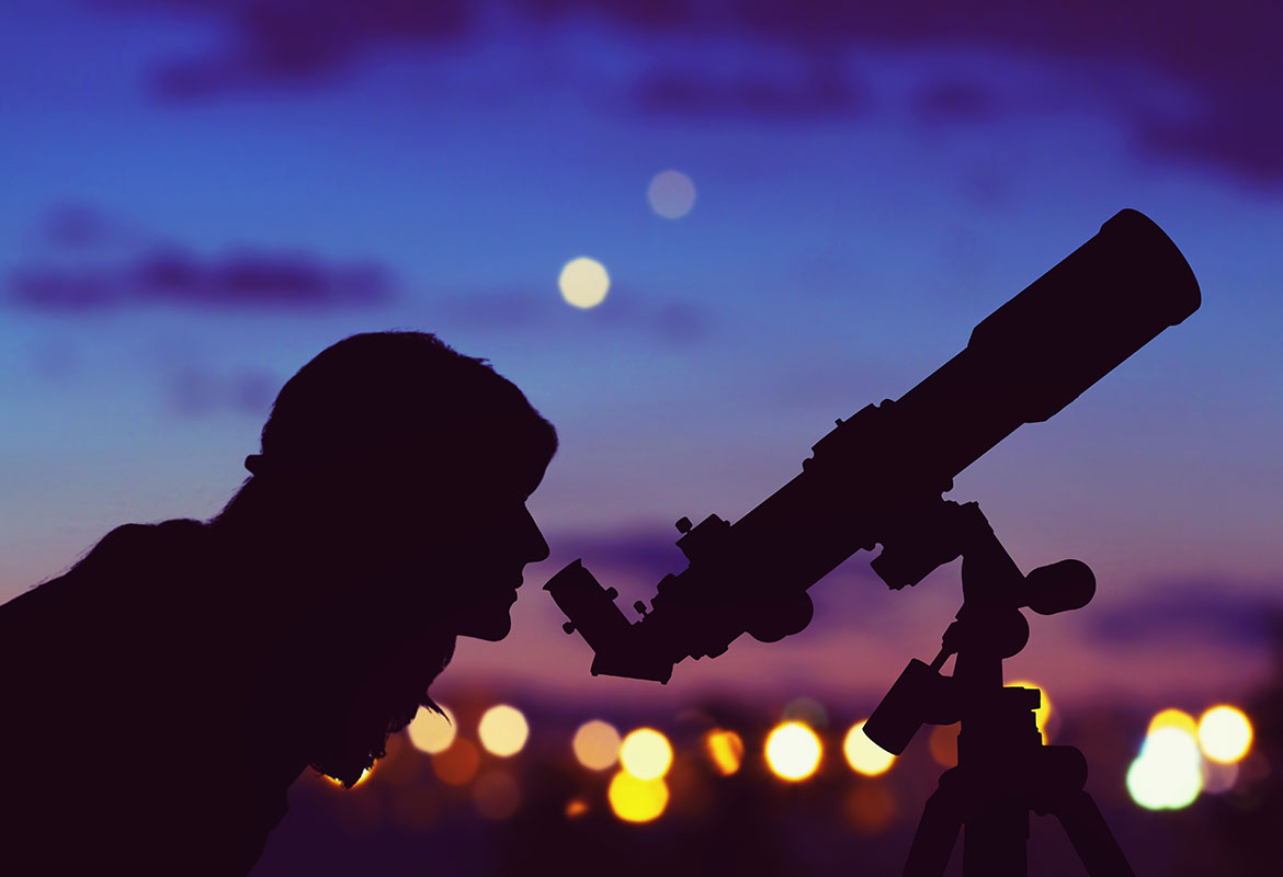 Silhouetted person looking through a telescope at dusk with city lights in the background