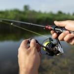Close-up of hands adjusting a spinning fishing reel on a rod by a calm lake with green vegetation in the background