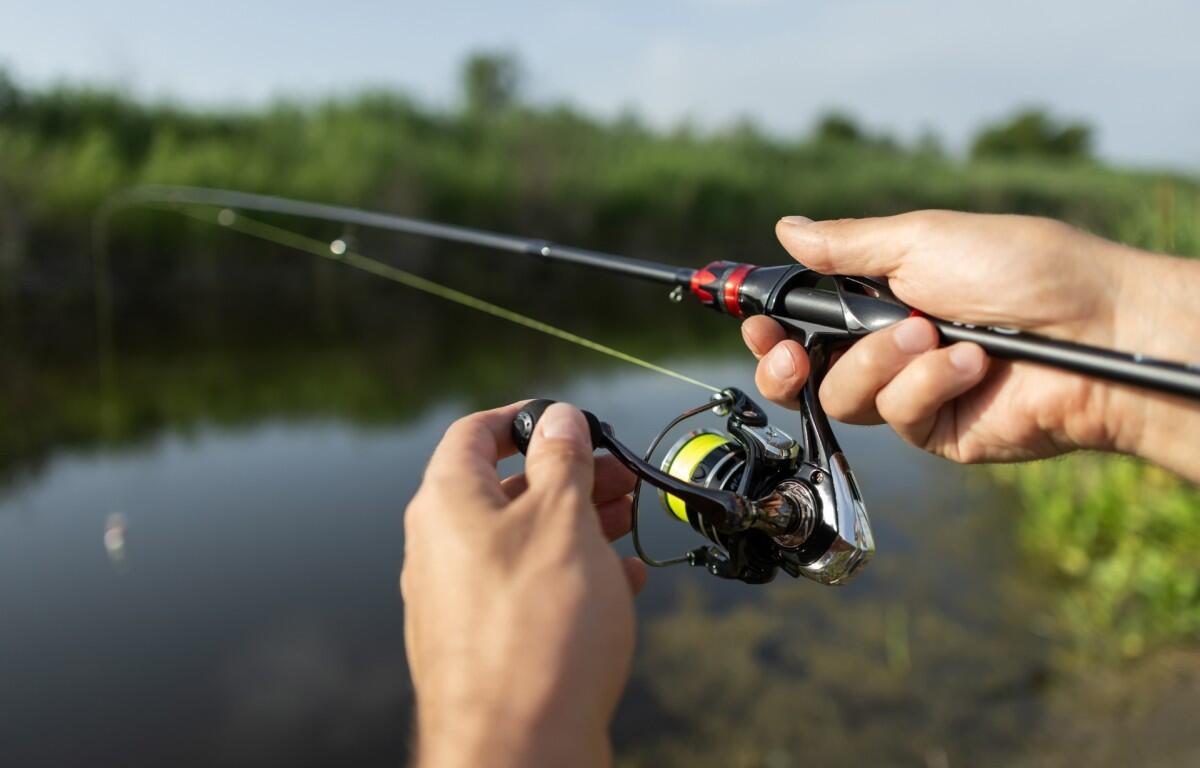 Close-up of hands adjusting a spinning fishing reel on a rod by a calm lake with green vegetation in the background