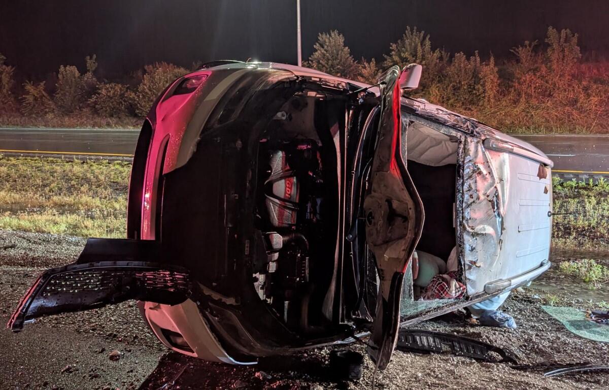 Red SUV flipped onto its side at night, door open and interior exposed amid debris on a roadside shoulder