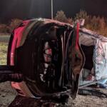 Red SUV flipped onto its side at night, door open and interior exposed amid debris on a roadside shoulder