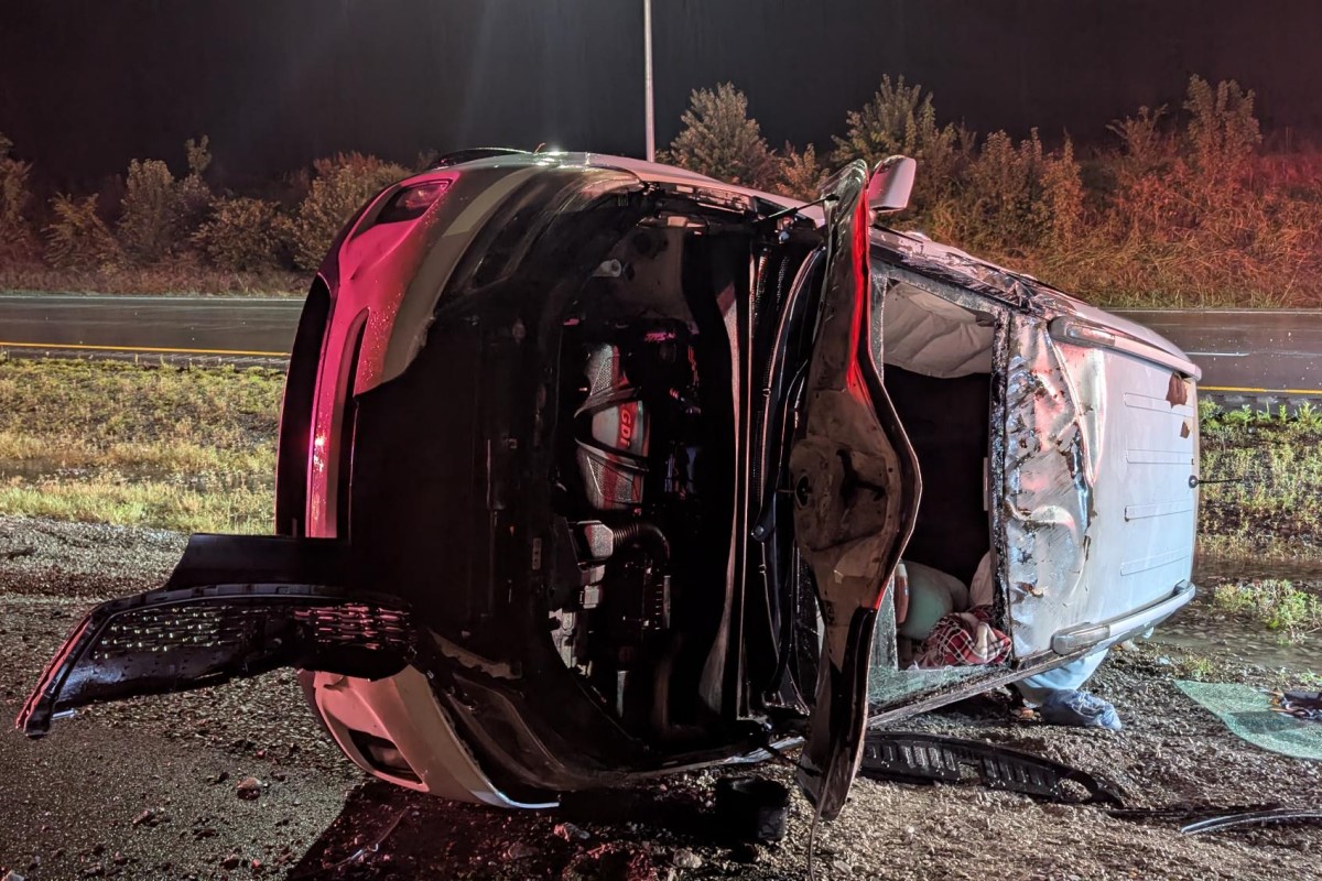 Red SUV flipped onto its side at night, door open and interior exposed amid debris on a roadside shoulder