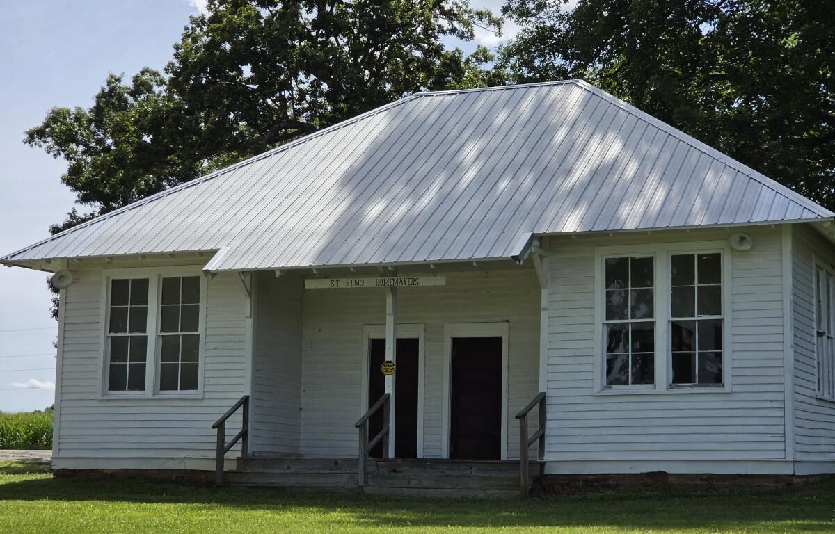 White wooden community building with a tall, silver metal roof, two front doors, and a small porch with railings under a sign that reads 'St. Elmo Homemakers'.