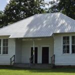 White wooden community building with a tall, silver metal roof, two front doors, and a small porch with railings under a sign that reads 'St. Elmo Homemakers'.
