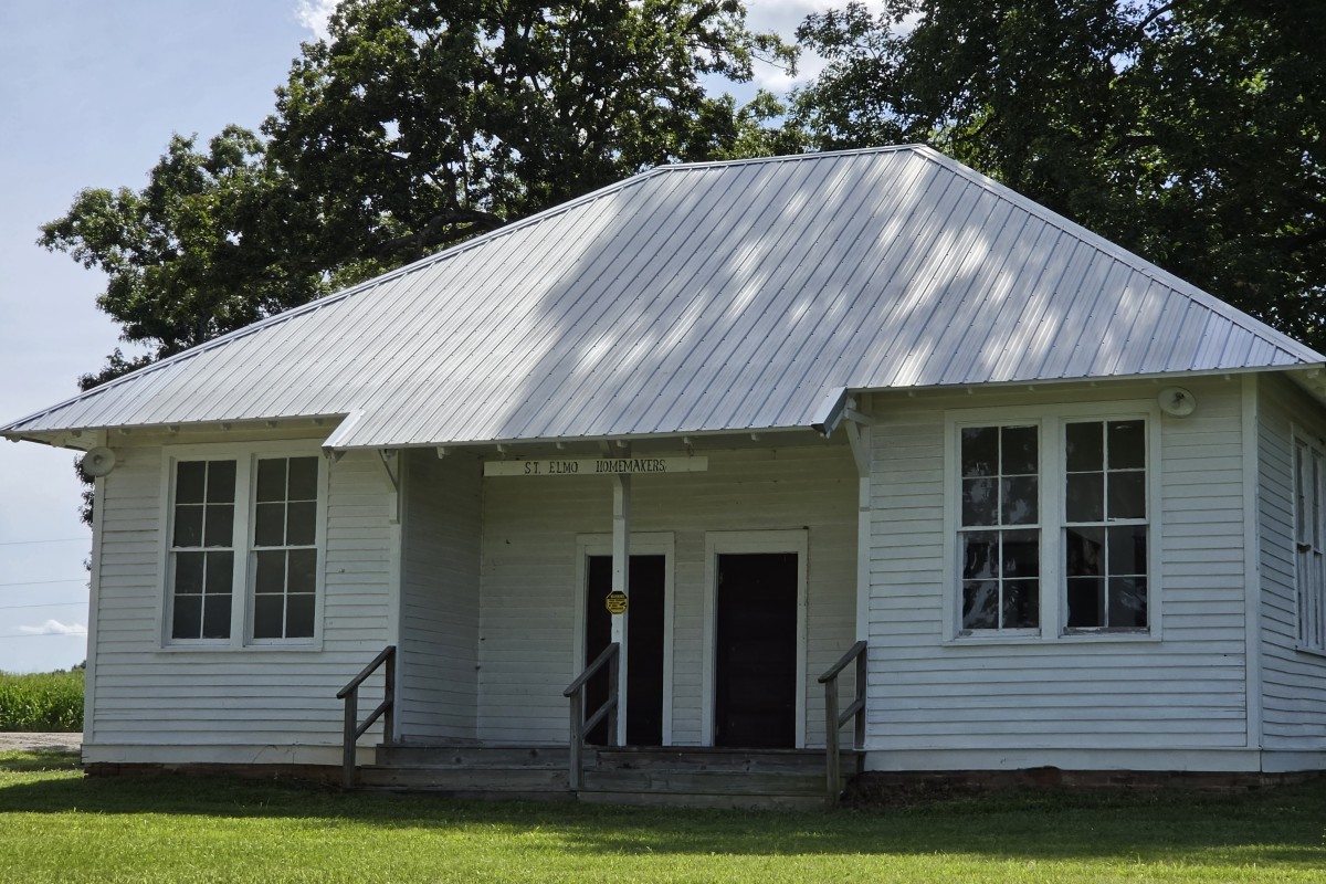 White wooden community building with a tall, silver metal roof, two front doors, and a small porch with railings under a sign that reads 'St. Elmo Homemakers'.