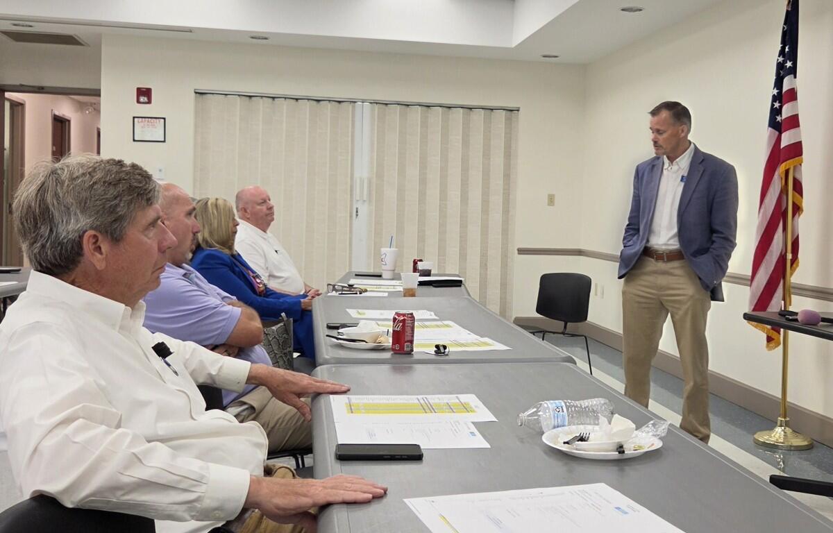 A man in a blue blazer speaks to a row of seated adults in a conference room, with an American flag in the corner and tables filled with papers and drinks.