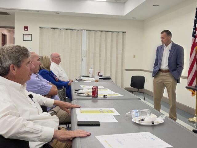 A man in a blue blazer speaks to a row of seated adults in a conference room, with an American flag in the corner and tables filled with papers and drinks.