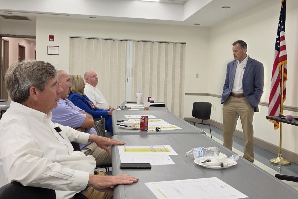 A man in a blue blazer speaks to a row of seated adults in a conference room, with an American flag in the corner and tables filled with papers and drinks.