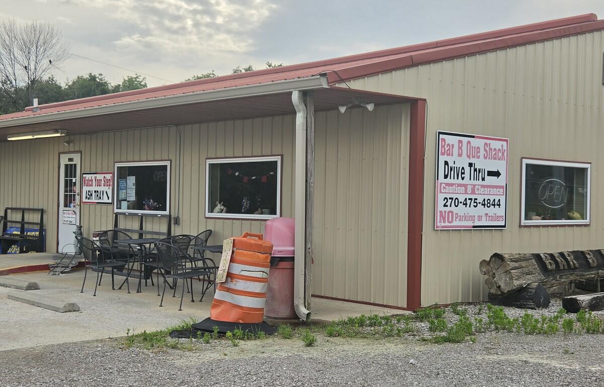 Small beige building with red trim and a large Bar B Que Shack sign, Drive Thru →, and outdoor seating under a covered area.