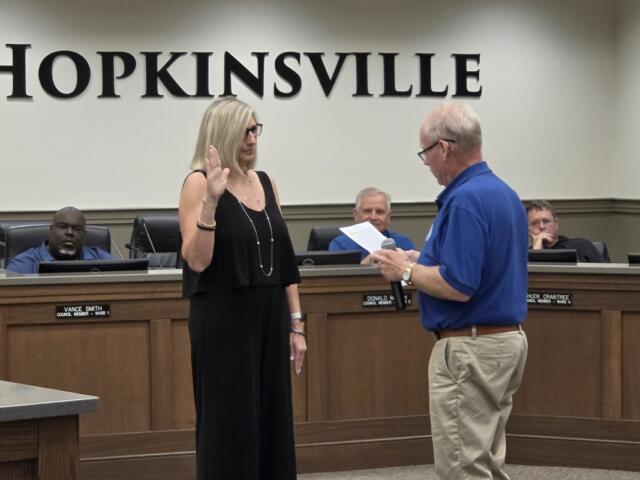 City council chamber scene with a woman raising her right hand as a man reads from a document for an oath ceremony in Hopkinsville courtroom setting