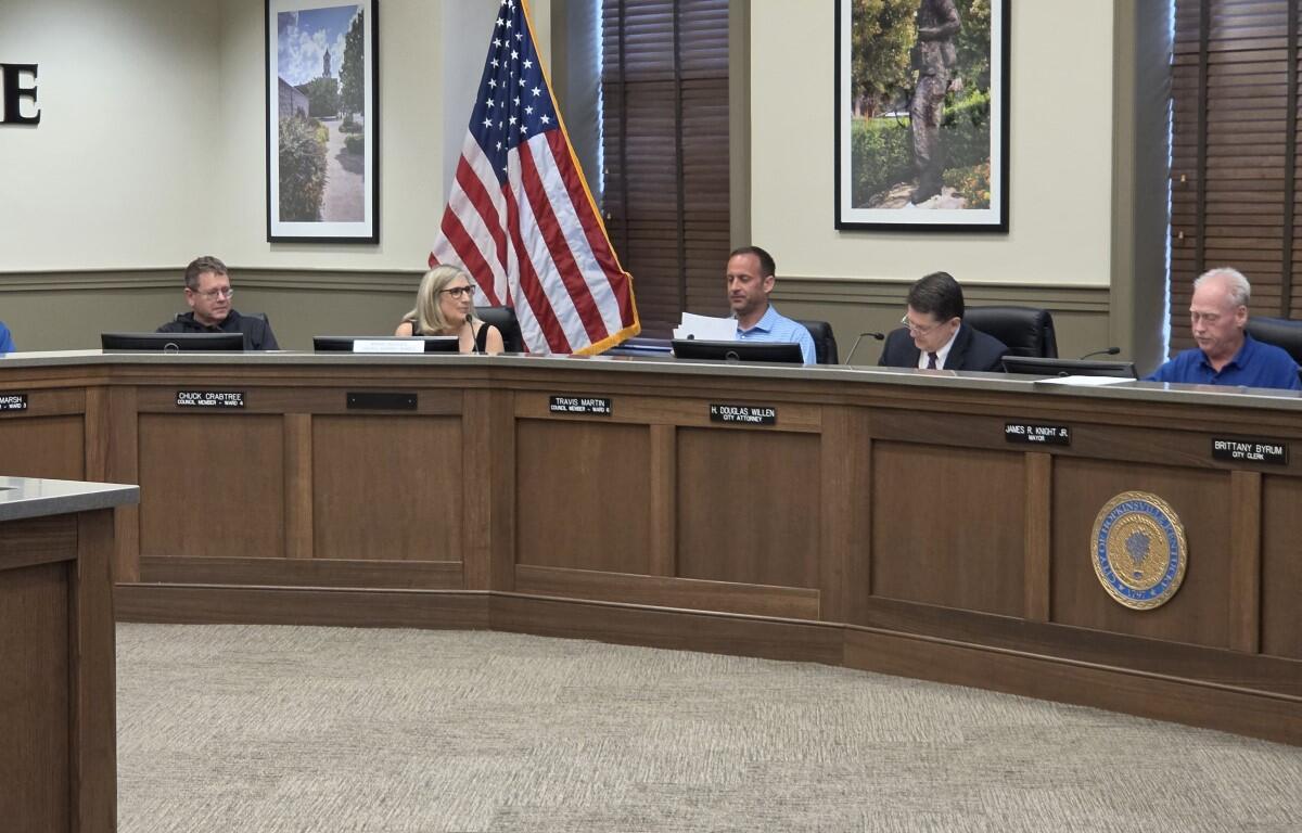 Council members seated at a curved wooden dais in a meeting room, with an American flag, framed photos, and a city seal visible on the front panel.