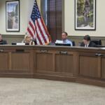 Council members seated at a curved wooden dais in a meeting room, with an American flag, framed photos, and a city seal visible on the front panel.