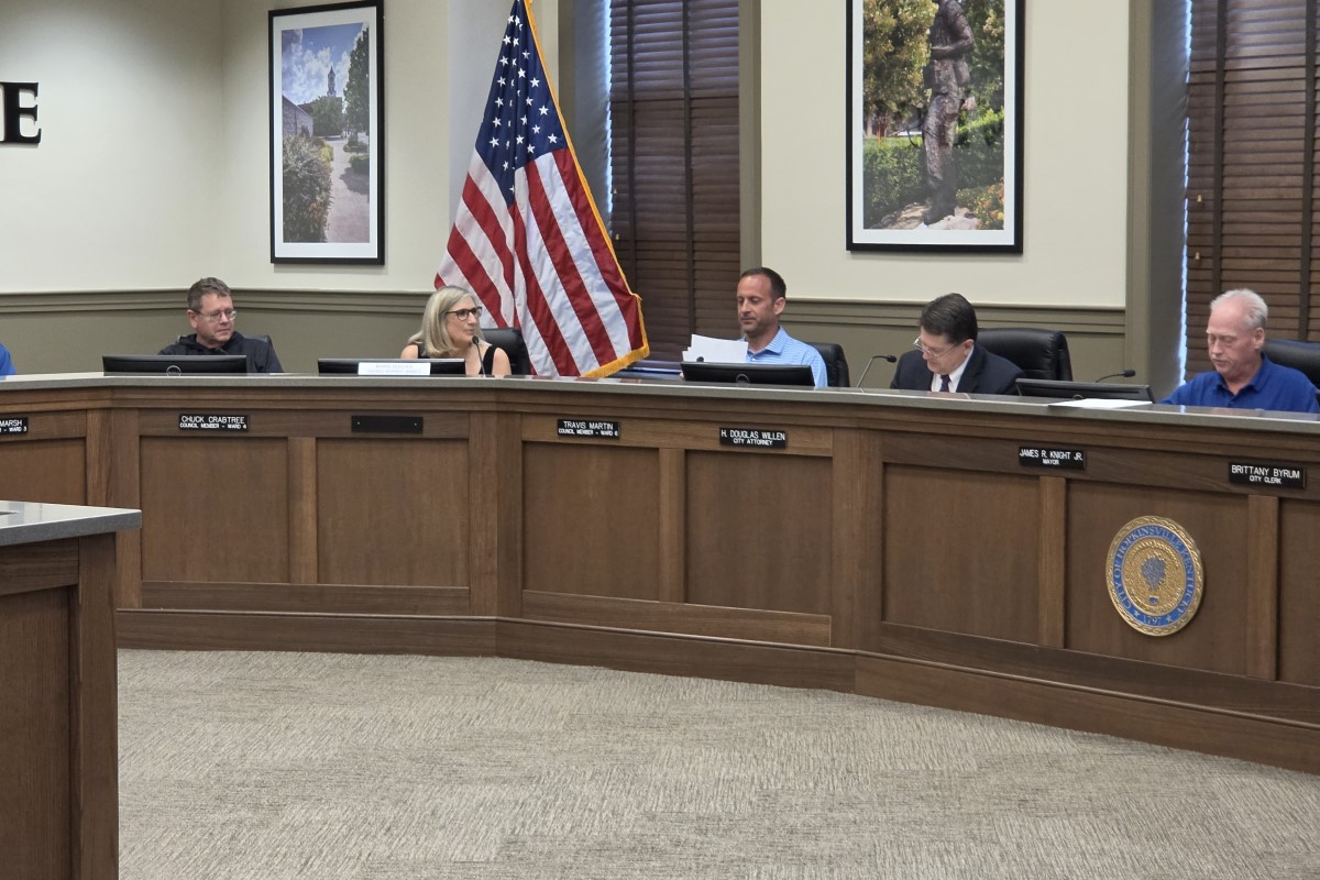 Council members seated at a curved wooden dais in a meeting room, with an American flag, framed photos, and a city seal visible on the front panel.
