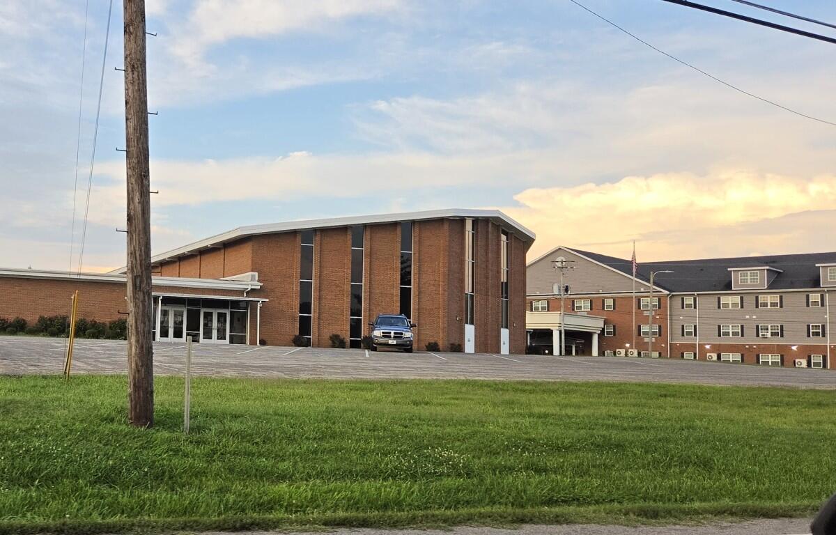 Brick institutional building with tall windows and a small entrance canopy, a blue SUV parked near the front, green lawn in the foreground, and power lines crossing the sky at dusk.