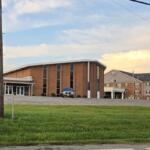 Brick institutional building with tall windows and a small entrance canopy, a blue SUV parked near the front, green lawn in the foreground, and power lines crossing the sky at dusk.