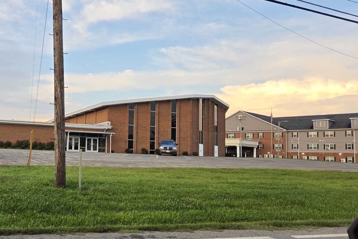 Brick institutional building with tall windows and a small entrance canopy, a blue SUV parked near the front, green lawn in the foreground, and power lines crossing the sky at dusk.