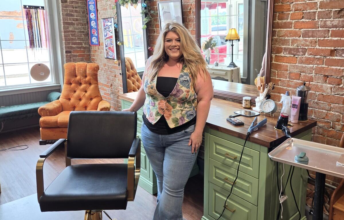 Woman with long blond hair standing in a hair salon, smiling, beside a green counter with styling tools and brick wall backdrop
