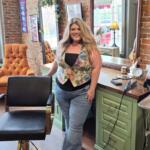 Woman with long blond hair standing in a hair salon, smiling, beside a green counter with styling tools and brick wall backdrop