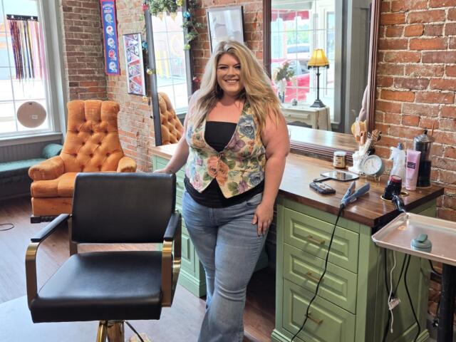 Woman with long blond hair standing in a hair salon, smiling, beside a green counter with styling tools and brick wall backdrop