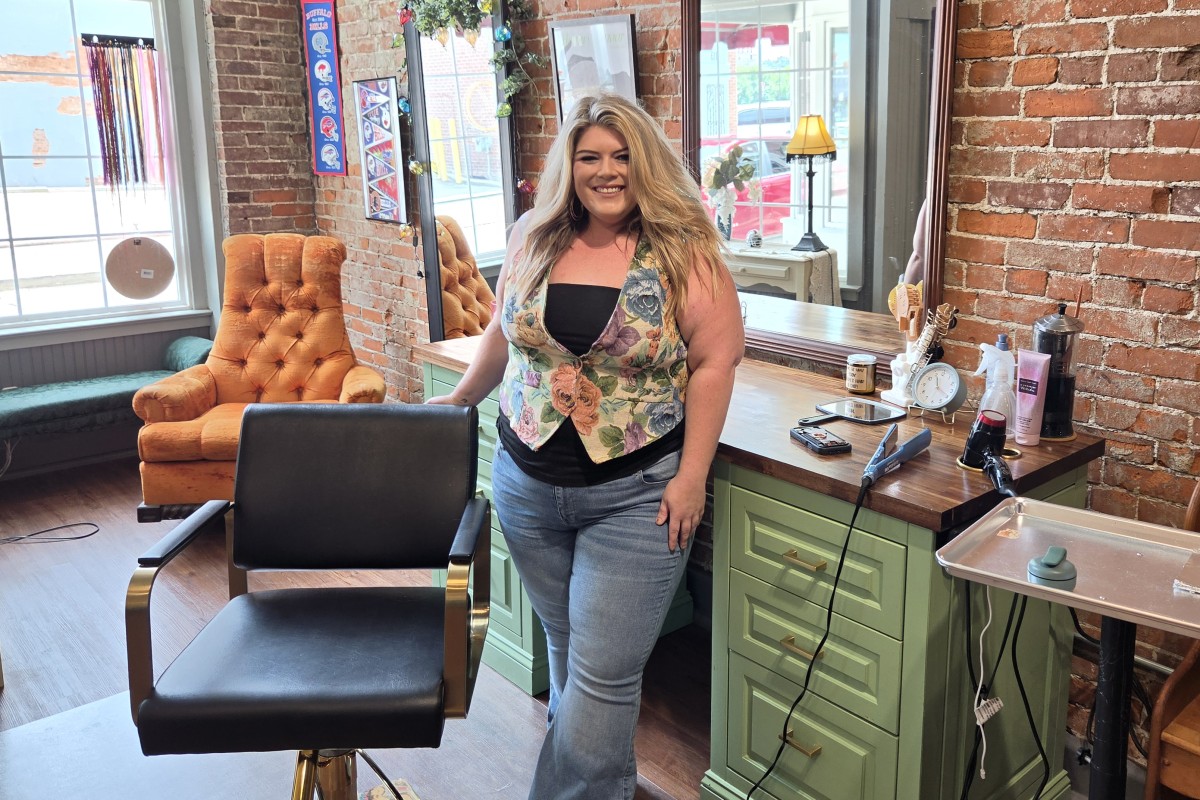 Woman with long blond hair standing in a hair salon, smiling, beside a green counter with styling tools and brick wall backdrop