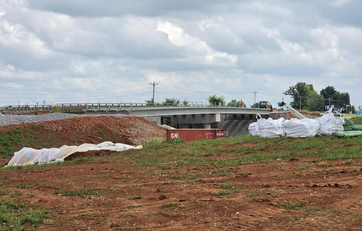 Construction site showing a bridge under construction, dirt piles, and large white geotextile bags on the ground beneath a cloudy sky.