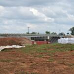 Construction site showing a bridge under construction, dirt piles, and large white geotextile bags on the ground beneath a cloudy sky.