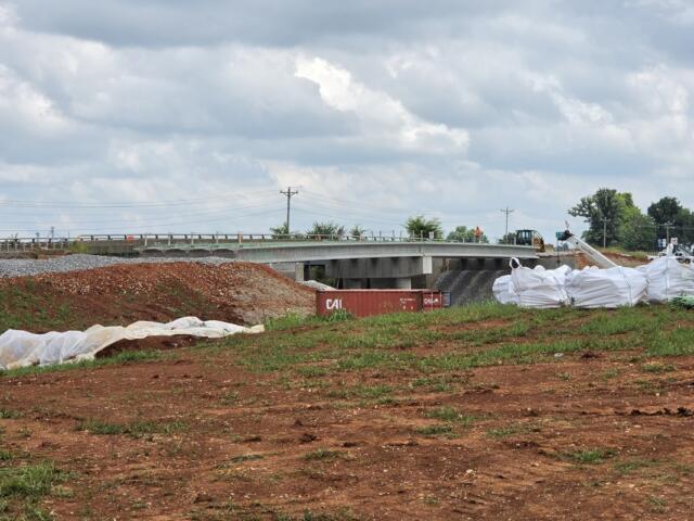 Construction site showing a bridge under construction, dirt piles, and large white geotextile bags on the ground beneath a cloudy sky.