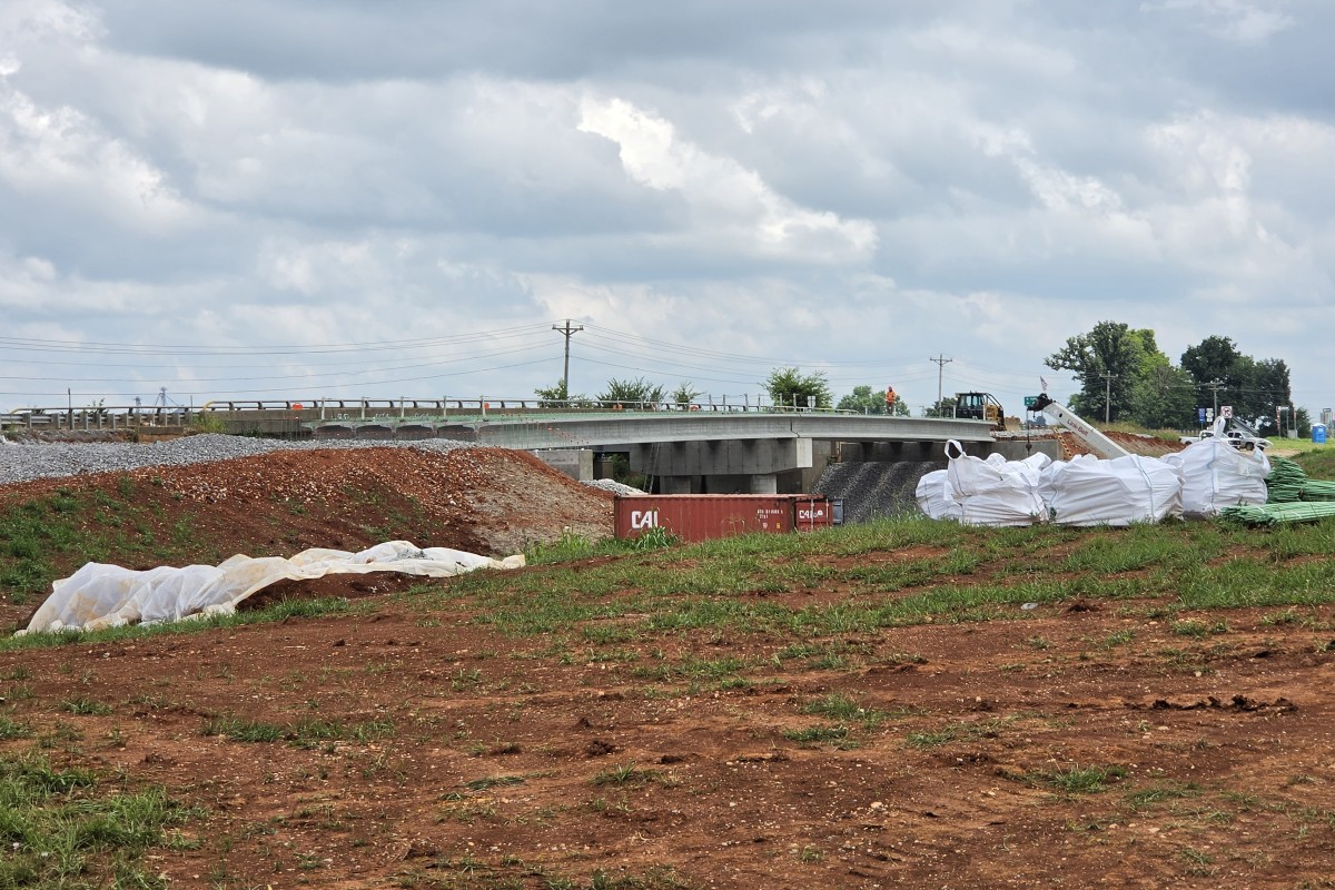 Construction site showing a bridge under construction, dirt piles, and large white geotextile bags on the ground beneath a cloudy sky.
