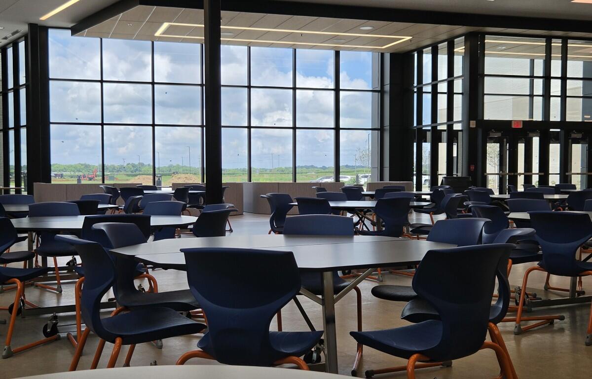 Bright indoor cafeteria with round tables and blue plastic chairs, large windows overlooking a green field and blue sky outside.