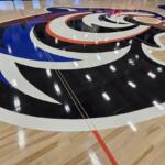 Basketball court floor with a large circular team logo painted at center in black, white, blue, and orange colors.
