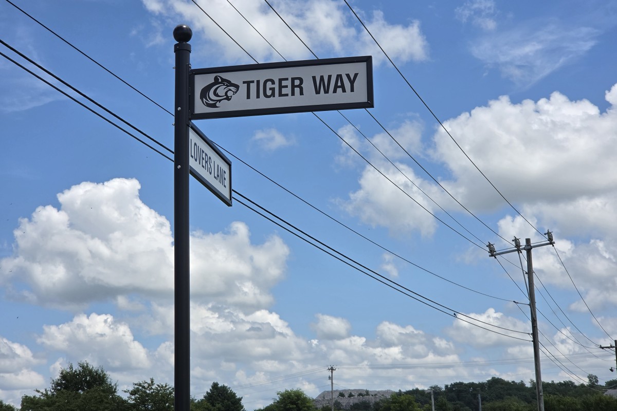 Street sign with 'Tiger Way' and a tiger logo, and a smaller 'Lovers Lane' sign on a tall black pole against a blue sky.