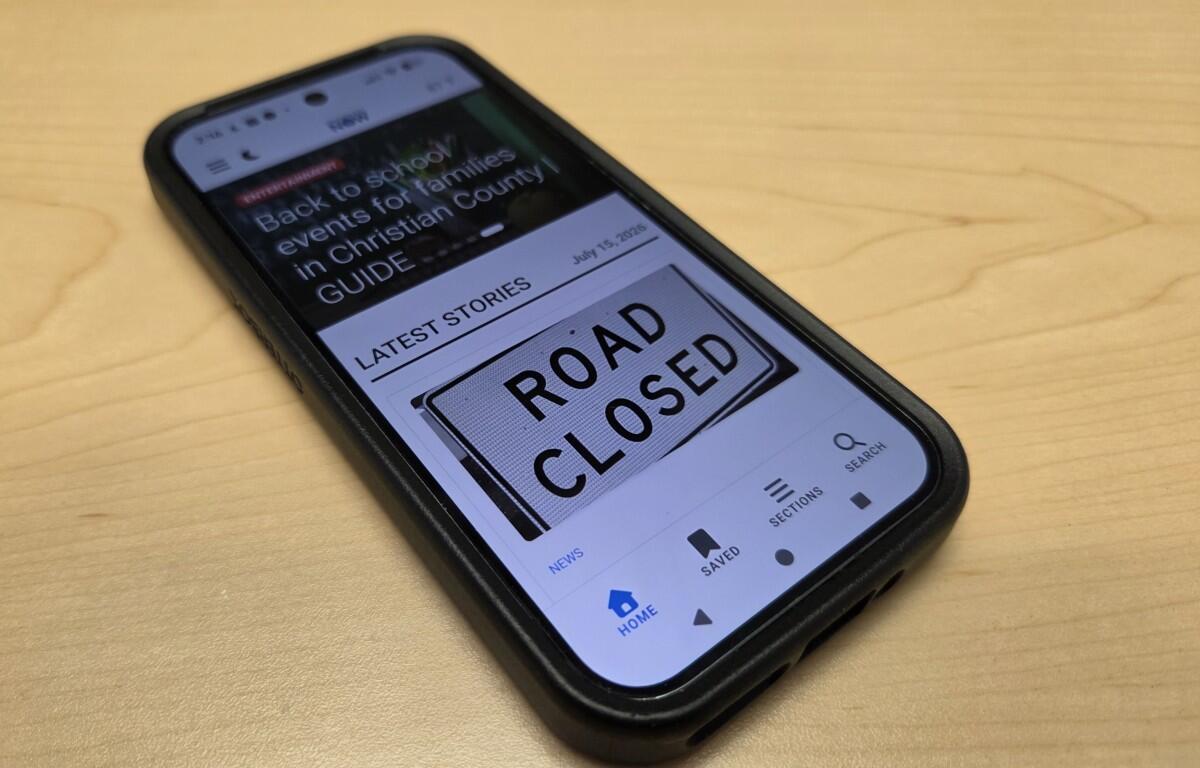 Smartphone on a wooden desk displaying a news app with a large 'ROAD CLOSED' sign on the screen, headline visible above the sign.