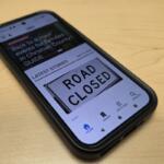 Smartphone on a wooden desk displaying a news app with a large 'ROAD CLOSED' sign on the screen, headline visible above the sign.