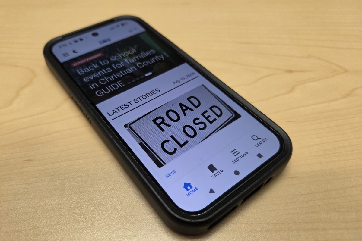 Smartphone on a wooden desk displaying a news app with a large 'ROAD CLOSED' sign on the screen, headline visible above the sign.