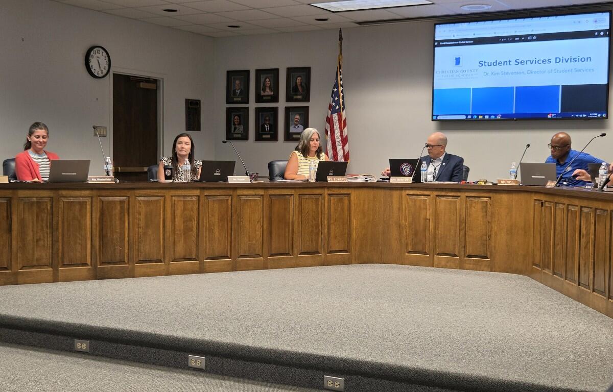 Panel of six officials seated behind a long wooden desk in a conference room, with microphones and laptops, US flag nearby and a large screen in the back showing 'Student Services Division'.
