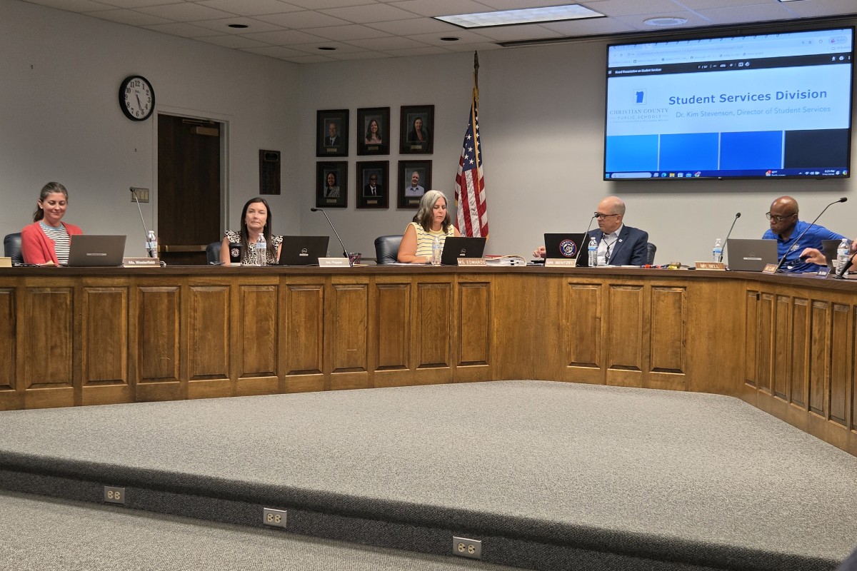 Panel of six officials seated behind a long wooden desk in a conference room, with microphones and laptops, US flag nearby and a large screen in the back showing 'Student Services Division'.