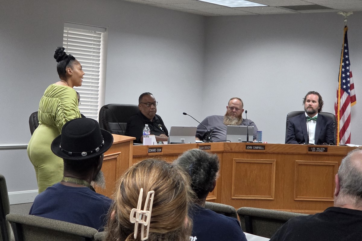 Woman in a lime-green dress speaks at a podium in a meeting room facing a panel of four men seated at a long desk, with an American flag in the corner and audience members in the foreground.