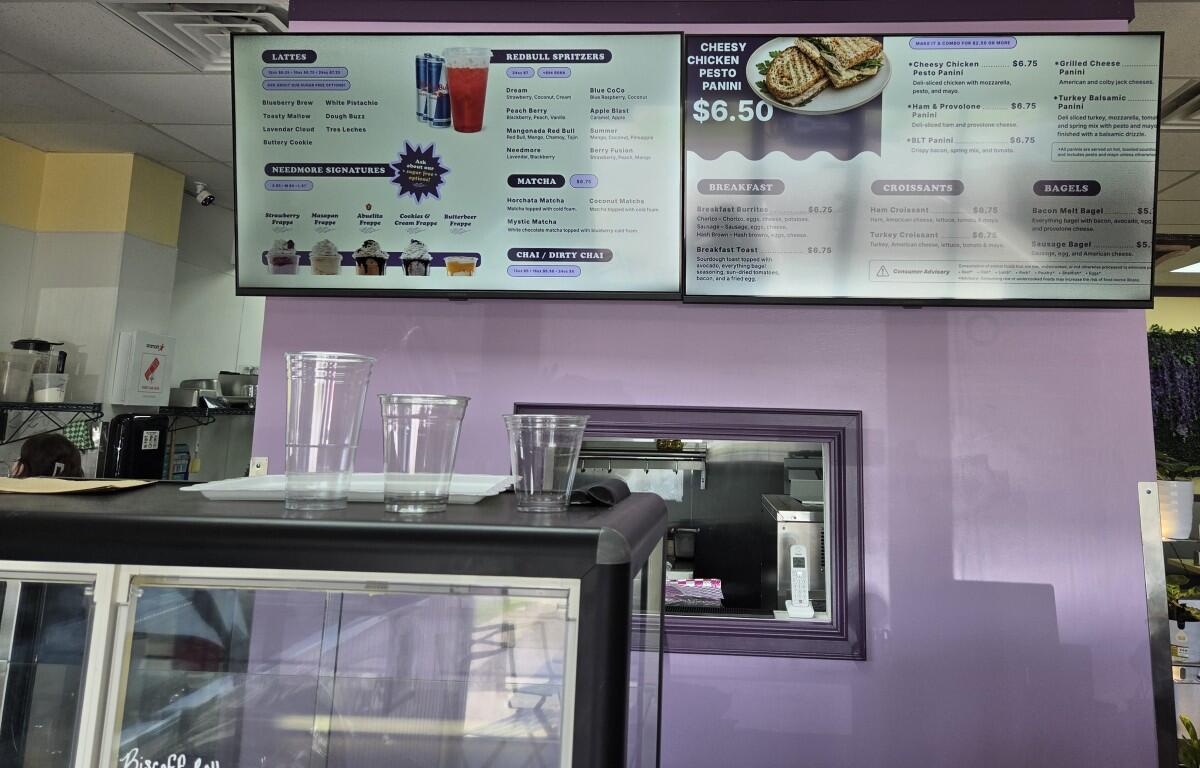 Cafe counter with a purple wall, two large menu boards overhead, several clear plastic cups on the counter, and a glass display case in front.