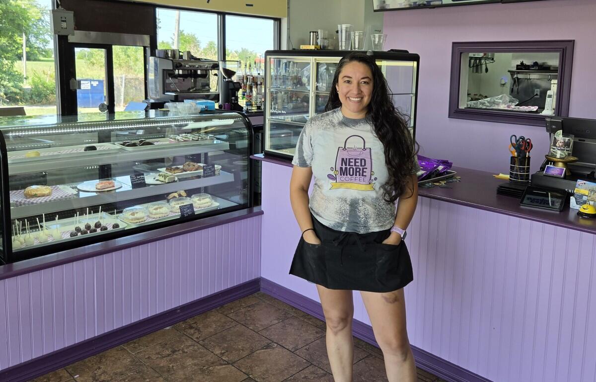 Smiling woman in a gray 'Need More Coffee' shirt stands behind a glass pastry display in a purple bakery/cafe.