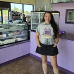Smiling woman in a gray 'Need More Coffee' shirt stands behind a glass pastry display in a purple bakery/cafe.