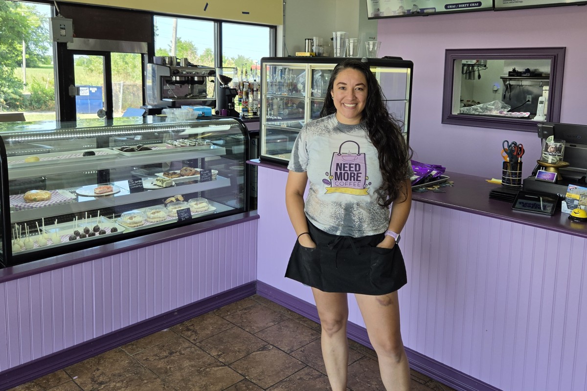 Smiling woman in a gray 'Need More Coffee' shirt stands behind a glass pastry display in a purple bakery/cafe.