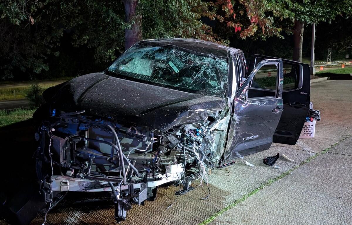 Black Toyota Tundra pickup severely damaged in a nighttime crash; hood crumpled, windshield shattered, doors open beside the road.