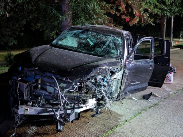 Black Toyota Tundra pickup severely damaged in a nighttime crash; hood crumpled, windshield shattered, doors open beside the road.