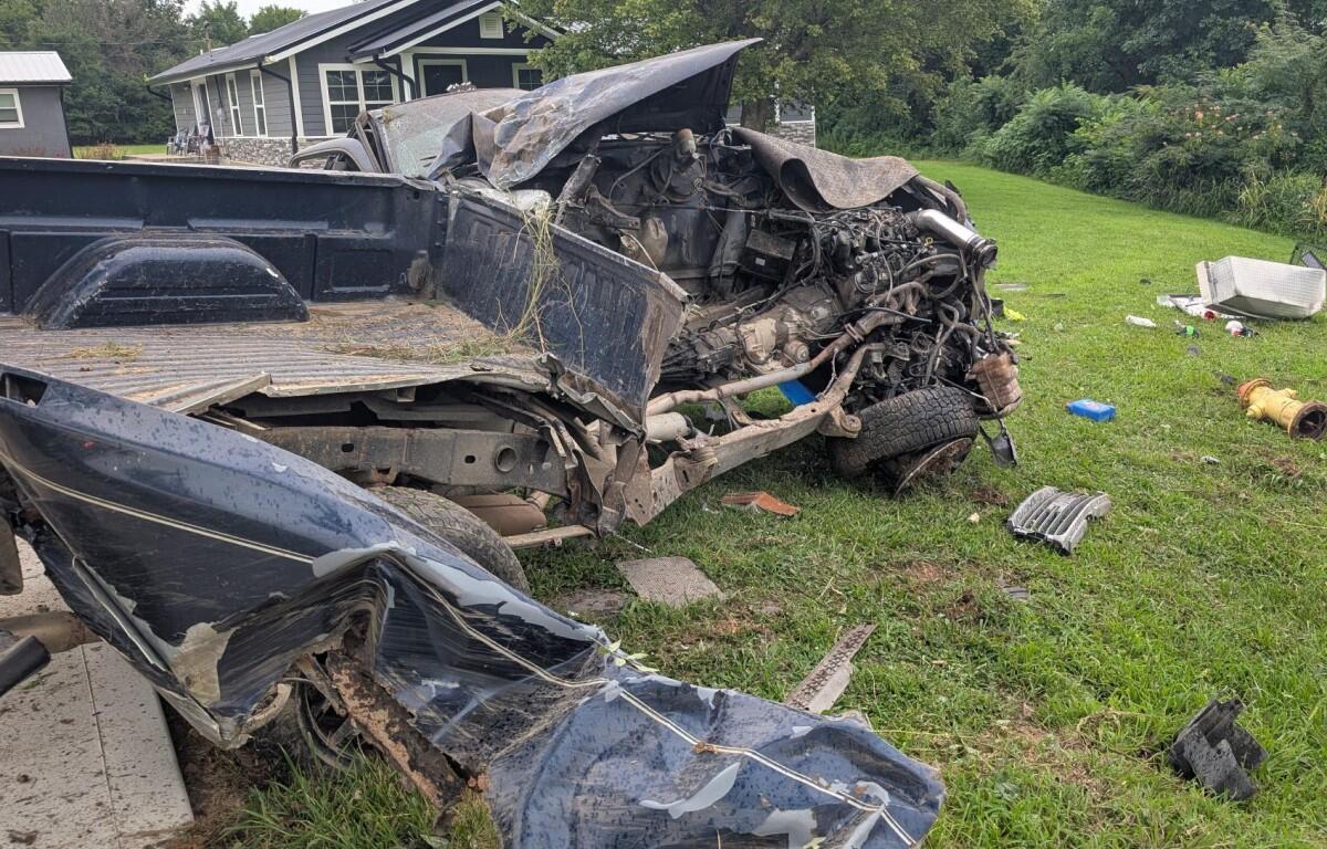 Dark blue pickup truck wreck with crushed front, exposed engine, and scattered debris on a grassy lawn.