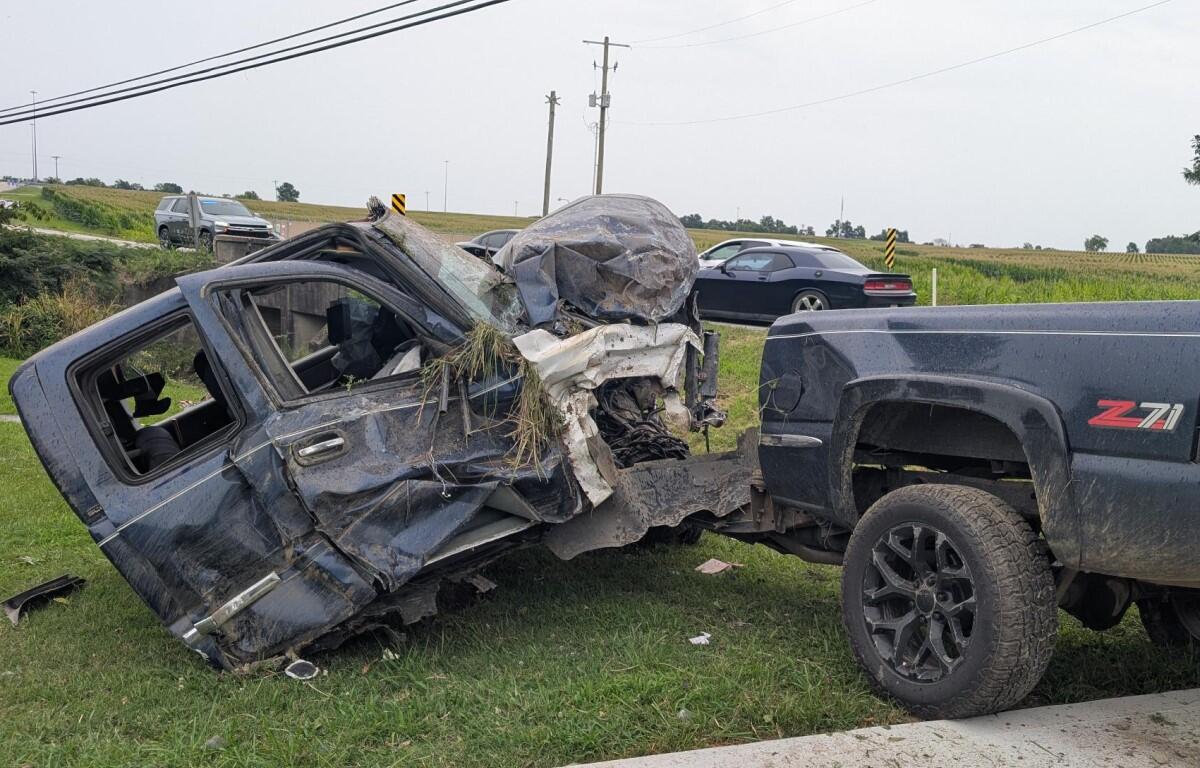 Pickup truck towing a severely damaged SUV on a grassy roadside after a collision, front end crushed and debris scattered nearby.