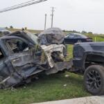 Pickup truck towing a severely damaged SUV on a grassy roadside after a collision, front end crushed and debris scattered nearby.