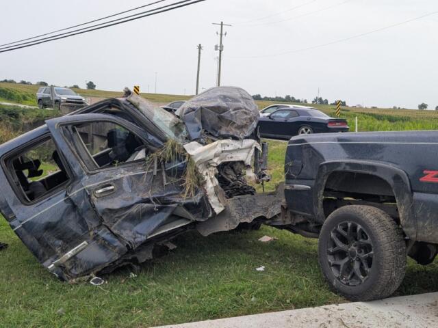 Pickup truck towing a severely damaged SUV on a grassy roadside after a collision, front end crushed and debris scattered nearby.