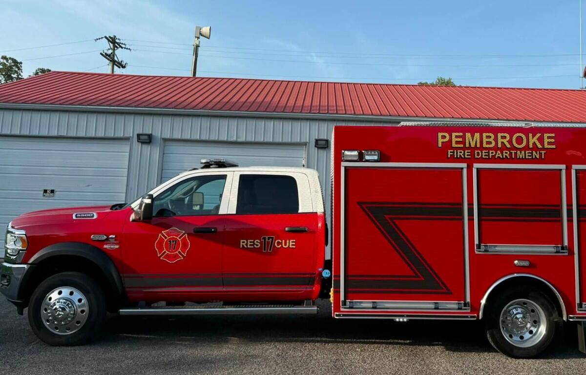 Red Pembroke Fire Department rescue truck parked outside a garage, with a blue sky above and a red roof building in the background.
