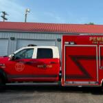 Red Pembroke Fire Department rescue truck parked outside a garage, with a blue sky above and a red roof building in the background.
