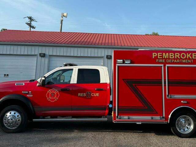 Red Pembroke Fire Department rescue truck parked outside a garage, with a blue sky above and a red roof building in the background.